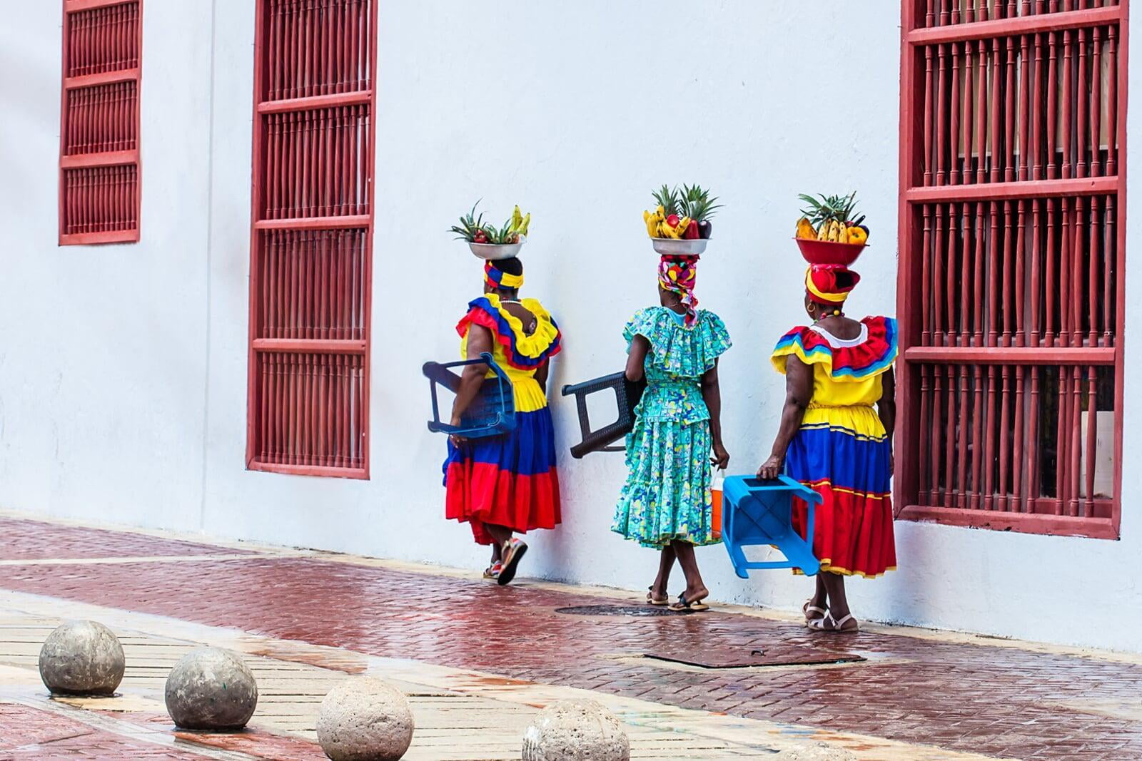 photo of colombian women with fruit on their head walking down the street with stools in their left hand